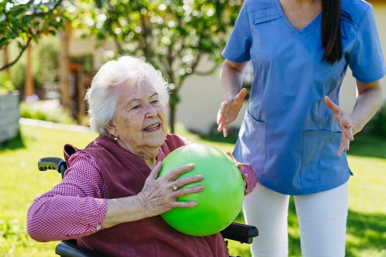 Two older adults seated at a small table with a daily activities calendar and a caregiver nearby.